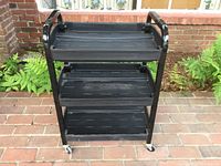 Front view of black rolling cart with three plastic shelves and black metal frame on brick surface with greenery in background.