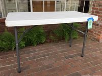 Full view of the folding table set up outdoors on a brick floor, showing the white plastic top and gray folding legs.