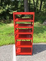 Front view of red six-shelf industrial metal shelving unit outdoors on pavement with greenery background.