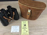 Binoculars alongside original brown leather carrying case with handwritten lot details.