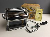 Full view of Marcato Atlas pasta machine made of chrome/plated metal, hand crank with black handle detached, original box behind.