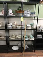 Full view of the Ikea metal and glass shelving unit containing various stacked plates to illustrate shelf usage. The shelving is black metal with glass shelves, tall and narrow, with a measuring stick for scale.