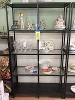 Full view of the black metal and glass shelving unit showing overall construction and shelving layout, also showing various household items on the shelves which are not included.