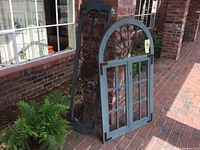 Two metal and wood wall art pieces leaning against brick, one arched and ornate with fleur de lis, one rectangular with window pane design.