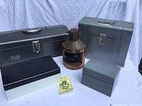 Wide shot of five metal boxes and a rusty lantern with blue dome, arranged on a white cloth background.