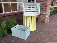 Three painted wooden crates stacked and arranged on outdoor brick surface near plants and window. Yellow crate largest, two smaller blue crates in front.