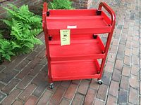 Full view of red utility cart with three tray shelves on caster wheels outdoors on brick surface.