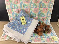 Two vintage baby quilts spread with one pastel cartoon-themed quilt in back and one folded blue floral-themed quilt in front, wooden alphabet blocks on right
