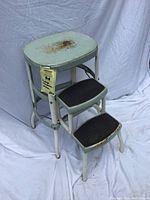 Front perspective of vintage metal step stool chair showing sea foam green top and two steps with black non-slip pads. Rust and paint stains visible.