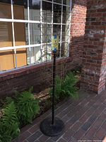 Full view of tall vintage metal postcard rack with rotating slots and heavy round base in outdoor setting with brick wall and plants.