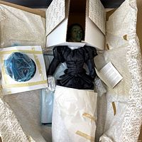 Porcelain doll wrapped in tissue and plastic inside the box with accessories and certificate visible