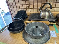 Photo of assorted bakeware including loaf pans stacked, Bundt cake pan in front, metal perforated baking trays below, stainless steel kettle on wire rack, and two wooden rolling pins behind