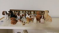 Photo showing 12 assorted ceramic dog figurines on a white shelf with a ceramic dog sign behind them