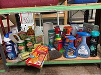 Various assorted cleaning and laundry supplies including bottles of disinfectants, cleaners, detergent, gloves, wipe containers, scrub pads, and broom head on green shelving.
