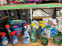 Overview of cleaning supplies on a wooden table, showing various liquid cleaners, sprays, powders, and paper towels.