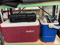 Two Coleman coolers stacked on shelf, black stove case on top of maroon cooler and metal stove partially seen in blue cooler.