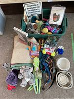 Green storage bin with various dog items inside including a plush mallard duck and pet boots, with other dog toys and accessories surrounding the bin.