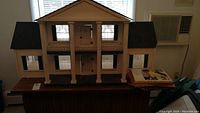 Front view of large wooden doll house with black roof and white pillars sitting on a table with doll house books stacked to the side.