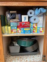 Kitchen supplies and paper products on shelf including various boxes of plastic wrap, foil, ziplock bags, paper towels, cleaning sponges, household wax paper, and steel wool pads along with some baking pans below.
