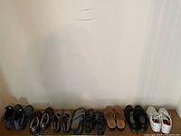 All nine pairs of ladies shoes arranged side by side on the floor against a wall, showing variety of styles and colors including sneakers and sandals.