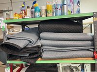 Shelf with black and gray quilted moving blankets, stacked and folded, with various cleaning products on the shelf above.