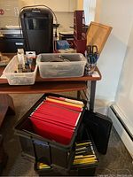 Photo of a Fellows paper shredder on a table behind two clear plastic containers with office supplies, a black mobile file box with red and yellow file folders, and various office tools like scissors and pens in cups.