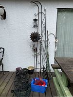 Outdoor setting with tall metal plant holders, metal garden stakes, decorative sunburst, black metal plant baskets, and a blue plastic bin with hummingbird feeder parts.
