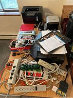 View of HP LaserJet and OfficeJet printers, Dell speaker subwoofer, various papers and electronics on table including power strips and cables.