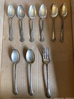 Top view showing six ornate demitasse spoons above two shell-handle teaspoons and one matching fork on cardboard.