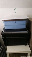 Stack of three plastic storage bins (light blue, black, black) placed on a small white table against a light wall.