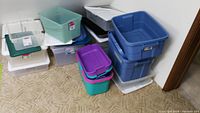 Group of various sized plastic storage bins in blue, purple, teal, and frosted white with lids nearby, stacked on carpet.