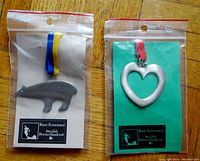 Two packaged pewter ornaments side by side on wood floor: polar bear on left with blue/yellow ribbon, heart on right with red ribbon