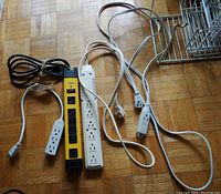 Four power strips with attached cords, varying lengths and outlets; one yellow and black NOMA power strip with six outlets and red switch, two white power strips with multiple outlets, and one smaller white power strip with three outlets and short cord.