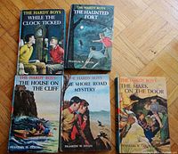 Photo showing front covers of all 5 Hardy Boys books with detailed cover illustrations: The House On The Cliff, The Shore Road Mystery, While The Clock Ticks, The Mark On The Door, The Haunted Fort.