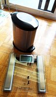 Bathroom garbage container and digital weighing scale placed on hardwood floor near a window. Shows the size and condition of both items.