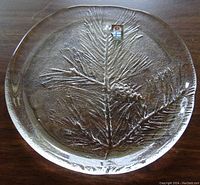 Clear round glass plate with embossed spruce branch motif and slight lip at edge, on wooden surface.