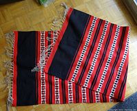Folded long woven mat displaying alternating horizontal bands of black, red, and white geometric pattern with fringed ends showing.
