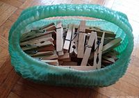 Light green textured plastic basket filled with about 70 wooden clothespins on hardwood floor