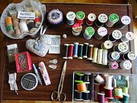 Overview photo of entire sewing lot including pin cushions, scissors, numerous spools of thread, needles, thread picker, and measuring tape arranged on wooden table