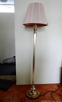Full view of the tall vintage Mid-Century Modern floor lamp standing against a wall, showing brass metal pole and beige pleated fabric lampshade.