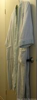 Front-angle view of the long white bathrobe hanging on a door, showing the belt, sleeves, and pockets with pale blue lining visible inside.