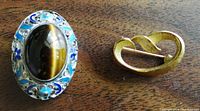 View of two brooches on a wooden surface, showing the detailed front of the silver-tone brooch with tiger's eye stone and the back of the gold-tone swirl brooch.