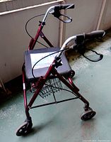 Side view of red metal frame rollator walker with black padded seat and mesh basket under seat, four caster wheels, hand brakes on handles. Missing back rest.