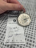 Photo of a hand holding a Gruen pocket watch on a checkered surface with a lot tag visible