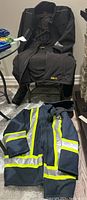 Two men's work coats displayed, one black Dewalt insulated coat with fleece lining hanging over chair, and one dark blue Work King coat with high visibility reflective stripes laid out below.