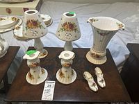 Photo showing two candle lanterns with pierced floral detailing, two candleholders with a similar floral pattern, the Old Country Roses vase with fluted top and pedestal base, and two shoe figurines all displaying the floral design and gold accents.
