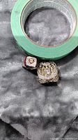 Two gold tone Phillies championship rings shown side-by-side on a grey cloth next to a green tape roll for scale.
