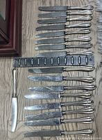 Top-down photo showing 17 silver butter knives in two different sizes, some knives stacked, placed on a wooden surface with measurement scale.