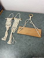 Three handmade macrame hangers laid out on a green surface. Two are macrame knotted hangers with fringes at bottom; the third is a wooden bar with hanging cotton cords and wooden ring.