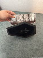 Black coffin-shaped crossbody bag with a cross engraved on the front and the product tag held above it.